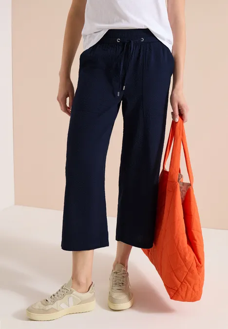 Frau in dunkelblauer Culotte, weißem Top, beigen Sneakern und orangener Tragetasche.