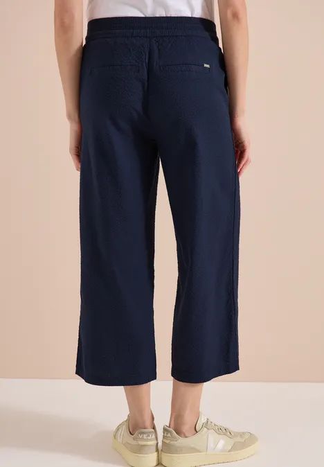 Rückenansicht: dunkelblaue, weite, knöchellange Hose mit Gummibund, Gesäßtasche und hellen Sneakers.