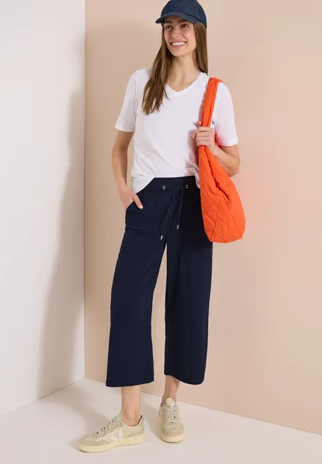 Frau mit weißem T-Shirt, weiten blauen Culottes, beigen Sneakern, orangener Tasche und Baseballcap.