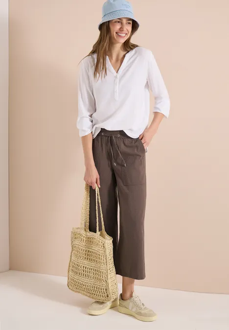 Frau trägt weiße Bluse, braune Culottes, beige Sneakers, Strohtasche und Hut.