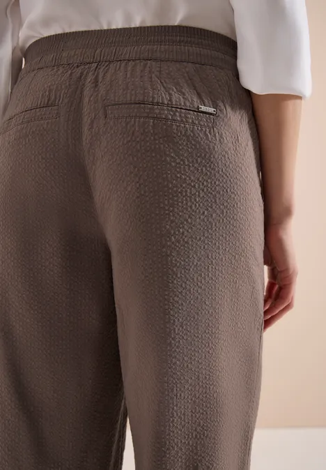 Rückenansicht einer Person in taupefarbener, texturierter Hose mit elastischem Bund und hinterer Reißverschlusstasche.