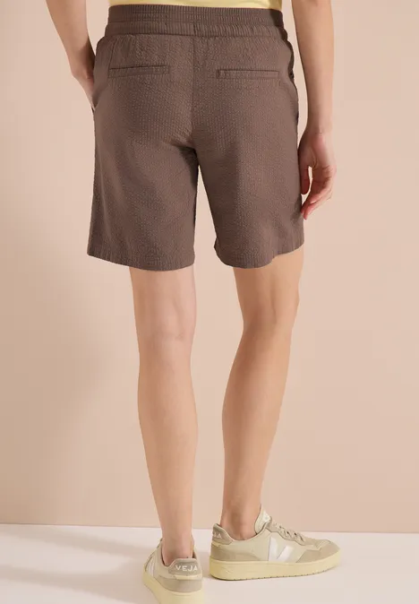 Rückenansicht einer Person in braunen Shorts und beigen Sneakers auf hellem Boden.
