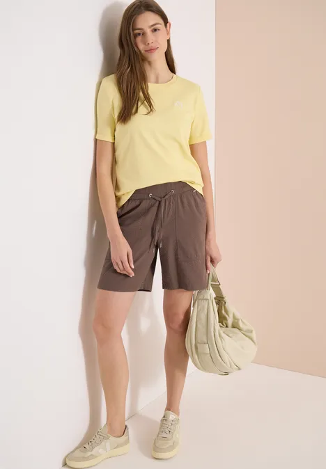 Frau in gelbem T-Shirt und braunen Shorts, trägt eine helle Tasche.