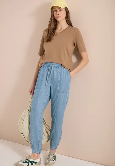 Frau in beigem T-Shirt, blauen Jogginghosen und Sneakers, hält eine große Tasche.