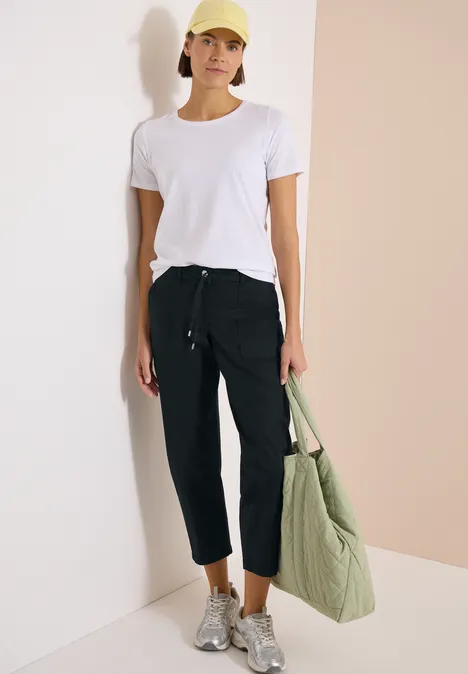 Frau trägt weißes T-Shirt, schwarze Knöchelhose, graue Sneakers und hält eine grüne, gesteppte Tasche.