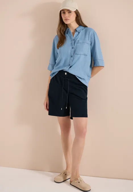 Person in einem hellblauen Hemd, navyblaue Shorts und beige Slipper vor neutralem Hintergrund.