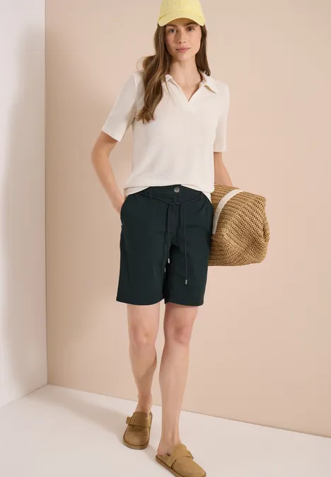 Frau in weißem Poloshirt, schwarzen Bermudashorts, braunen Loafers und Strohtasche.