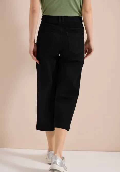 Frau steht rücklings in schwarzer, enkellanger Jeanshose und weißen Sneakern vor hellem Hintergrund.