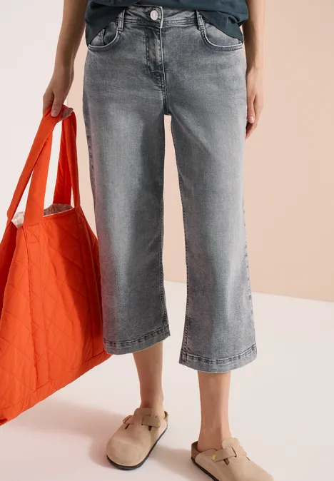 Wijde grijze 7/8-jeans, oranje tas in de hand en beige sloffen tegen een lichte achtergrond.
