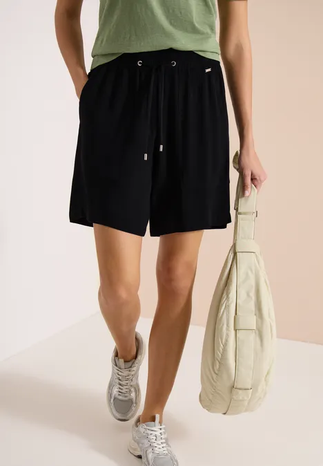 Frau in schwarzen Shorts, grünem T-Shirt, grauen Sneakern und großer beiger Tasche.