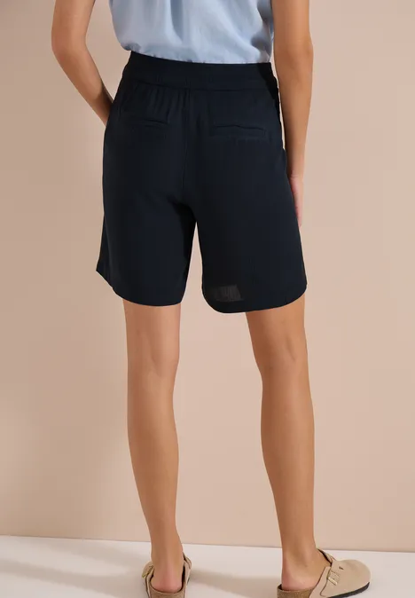 Rückenansicht einer Person mit hellblauem Oberteil und dunklen Shorts vor beigem Hintergrund