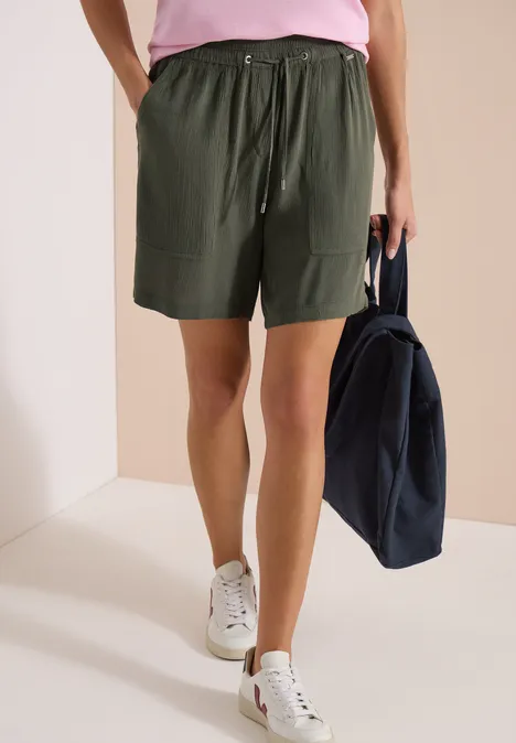 Frau in grünen Shorts, pinkem Shirt und weißen Sneakern mit einer dunkelblauen Tasche.