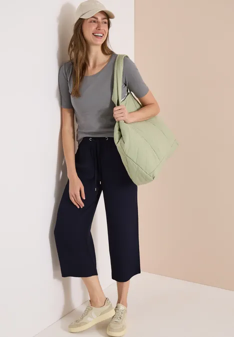 Frau mit grauem T-Shirt, dunkelblauen Culottes, beigen Sneakern und großer pastellgrüner Tasche vor hellem Hintergrund.
