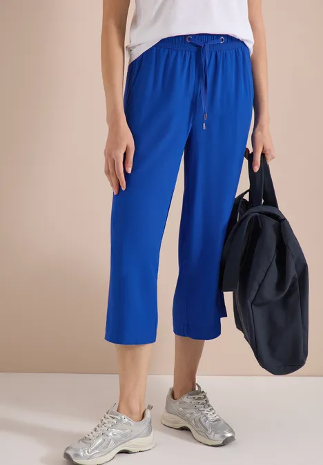 Person trägt blaue 7/8-Jogginghose, weißes Top, graue Sneaker und hält eine blaue Sporttasche.