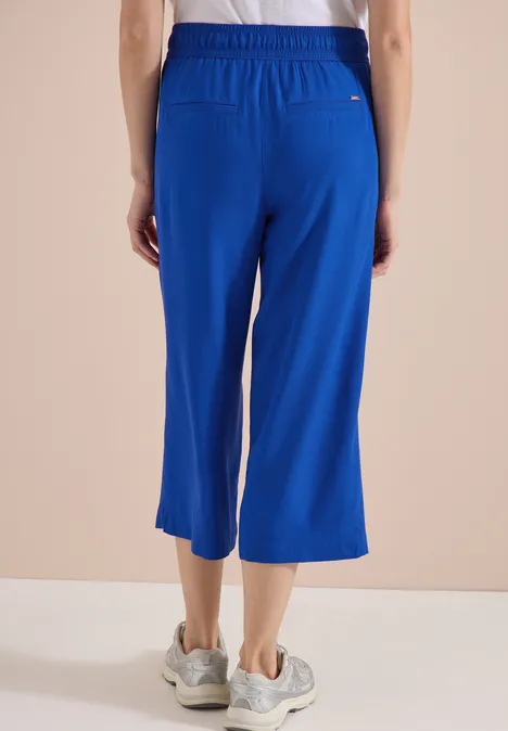 Person trägt weite, kniehohe blaue Hose mit elastischem Bund und weißen Sneakers vor beige Hintergrund.