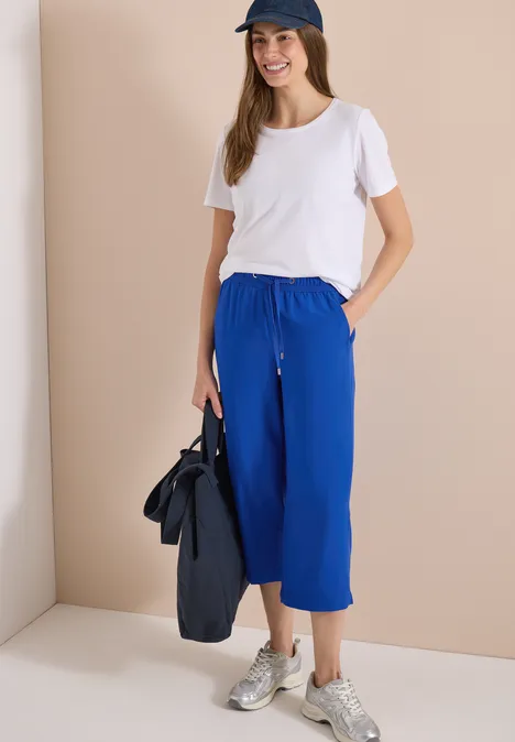 Frau in weißem T-Shirt und blauen Culotte-Hosen mit Sneakers, hält Tasche vor beigem Hintergrund.