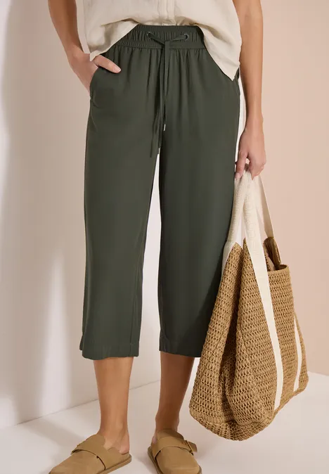 Damenmodell in olivgrüner Culotte mit Tunnelzug, weißem Top, Strohtasche und grünen Slip-ons