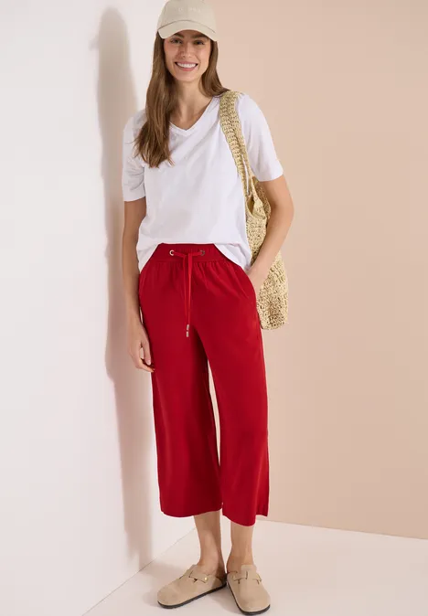 Frau lehnt an Wand, trägt weißes T-Shirt, rote Caprihose, Strohtasche, Espadrilles und Strohhut.