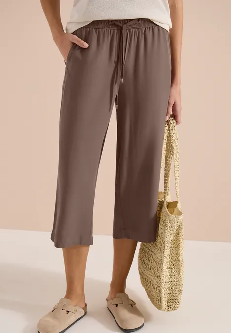 Braune, weite 3/4-Hose mit elastischem Bund, kombiniert mit beigen Slippern und geflochtener Strohtasche.