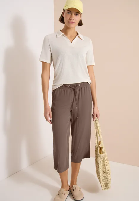 Frau trägt weißes Poloshirt und braune 7/8-Hose, hält geflochtene Einkaufstasche, lässiger Look.