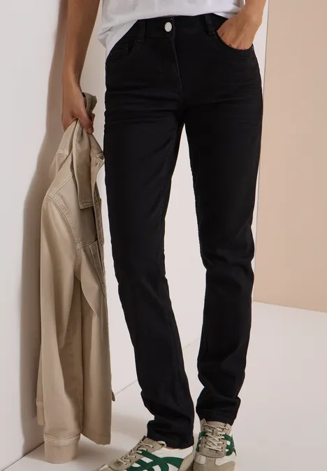 Schwarze, gerade geschnittene Jeans; Modell hält beigen Mantel und trägt gemusterte Sneaker