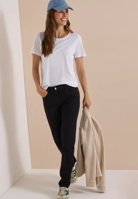 Frau in weißem T-Shirt, schwarzer Jeans und blauer Cap, hält eine beige Jacke in der Hand vor neutralem Hintergrund.