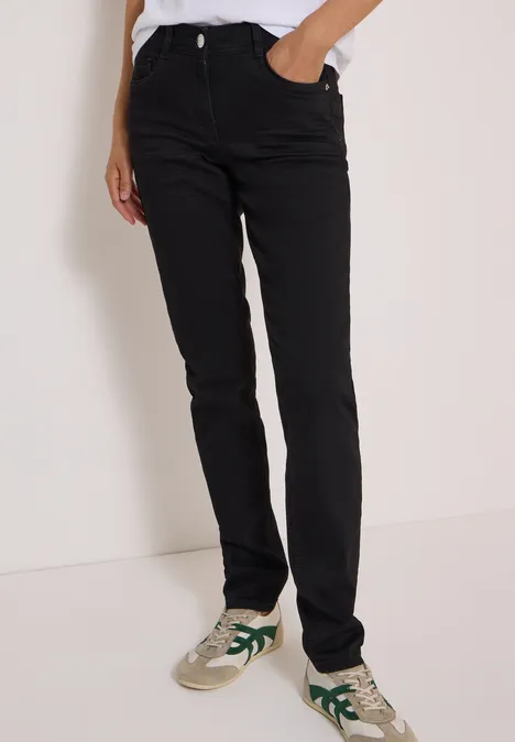 Schwarze gerade geschnittene Jeans mit weißen Sneakern mit grünen Details, Person steht vor neutralem Hintergrund.