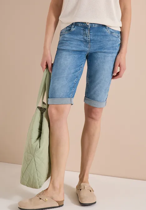Model trägt hellblaue Jeans-Bermudas, weiße Bluse, beige Loafers und eine hellgrüne gesteppte Tasche.