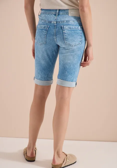 Frau von hinten in hellblauen, knielangen Jeans-Shorts mit Umschlag und beigen Loafern vor hellem Hintergrund.