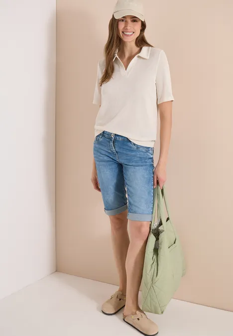Frau in hellem Top, Jeans-Bermuda und großer grüner Tasche vor beiger Wand.