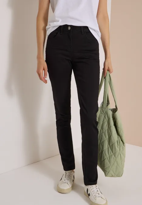 Jean taille haute Slim Leg, coupe slim Black Person trägt schwarze Slim-Fit-Hose, weißes T-Shirt, weiße Sneaker und hält eine grüne gesteppte Tasche.