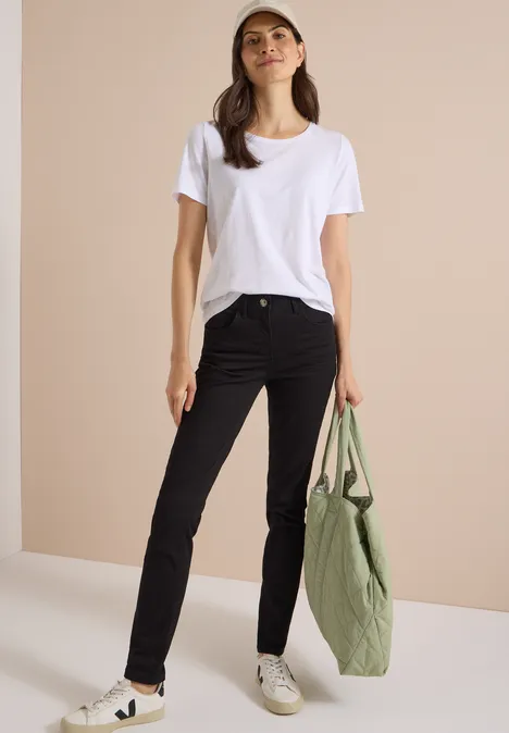 Jean taille haute Slim Leg, coupe slim Black Frau trägt weißes T-Shirt, schwarze Jeans und hält eine grüne Tasche.