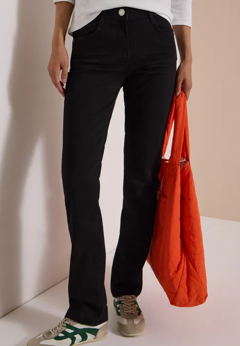 Schwarze Schlaghose, grün-weiße Sneaker, orangefarbene Steppjacke in der Hand.