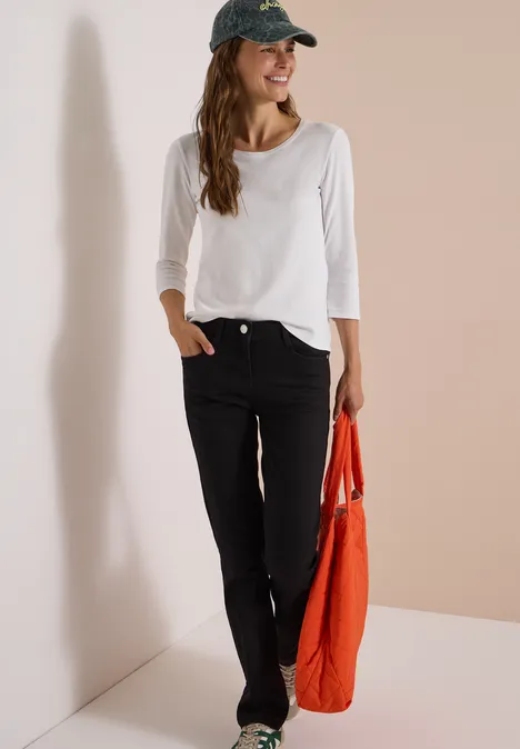 Frau in weißem Shirt und dunklen Jeans mit roter Tasche lehnt an Wand und lächelt