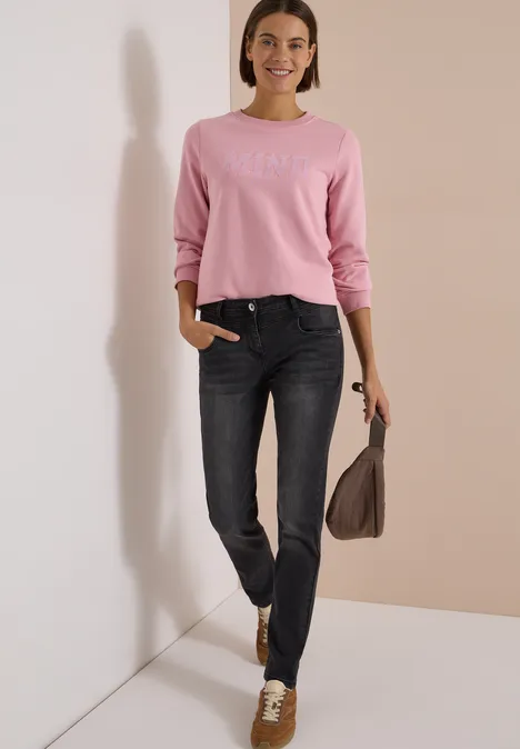 Een glimlachende vrouw in een roze trui en donker jeans houdt een bruine tas vast voor een lichte muur.