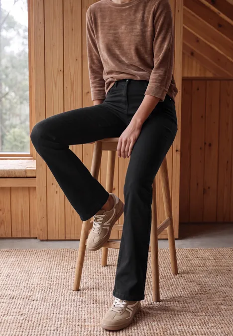 Eine Frau sitzt entspannt auf einem Holzschemel, trägt schwarze Jeans, braunen Pullover und helle Sneaker.