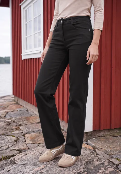 Frau trägt schwarze gerade Jeans und beige Schuhe vor rotem Haus am See.