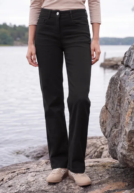 Eine Person steht am Ufer auf Felsen, trägt schwarze, gerade geschnittene Hose und beige Schuhe.