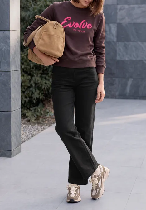 Vrouw met bruine trui, zwarte jeans en witte sneakers, draagt een tas.