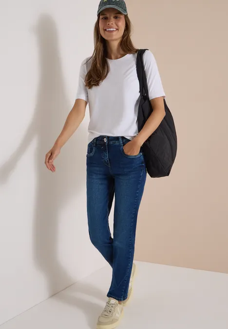 Jeans met High Waist en smalle pijpen in Slim Fit mid blue wash Lächelnde Frau in weißem T-Shirt, blauen Jeans, grüner Mütze und schwarzer Tasche vor heller Wand.
