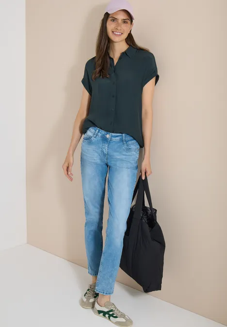 Mid Waist Slim Leg Jeans im Casual Fit light blue wash Frau in dunkelgrünem Oberteil, blauen Jeans, grünen Sneakern und einer schwarzen Tasche vor beigem Hintergrund.