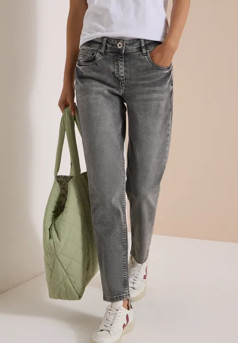 Mid Waist Slim Leg Jeans im Casual Fit light grey wash Person in grauen Jeans, weißem Shirt und weißen Sneakern trägt eine große grüne Stepp-Tasche.