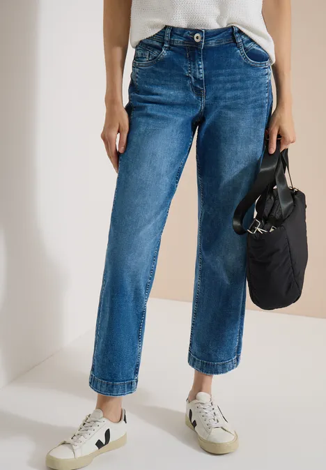 Vrouw draagt een licht gewassen, rechte, omgeslagen blauwe jeans met witte sneakers en een kleine zwarte tas.