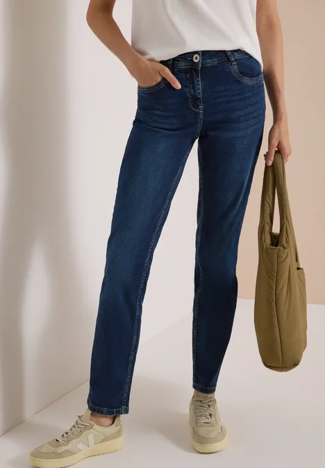 High Waist Straight Leg Jeans w kroju Slim Fit Dark blue wash Osoba ma na sobie ciemne niebieskie dżinsy, biały top i jasne trampki, trzyma zieloną torbę.