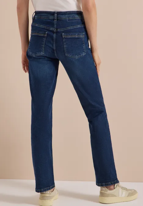 High Waist Straight Leg Jeans w kroju Slim Fit Dark blue wash Widok od tyłu osoby w ciemnoniebieskich, prostych jeansach i białych trampkach na beżowym tle.