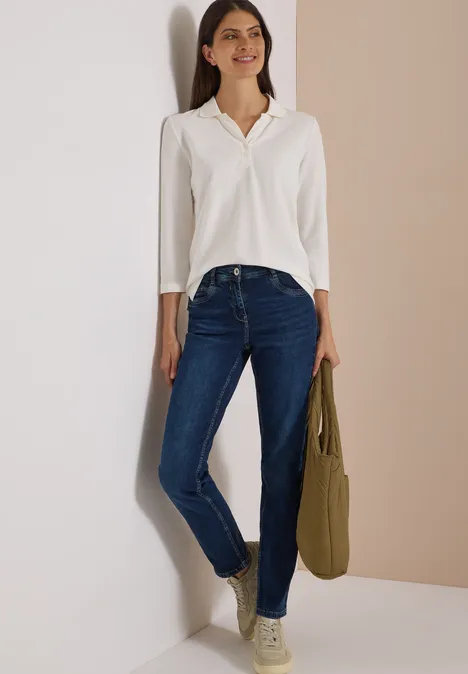 High Waist Straight Leg Jeans w kroju Slim Fit Dark blue wash Kobieta oparta o ścianę, uśmiechnięta, biała koszula, niebieskie dżinsy i oliwkowa torba.