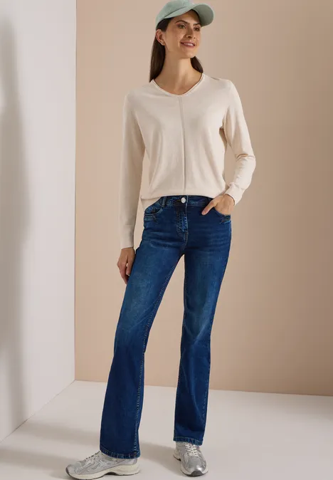 High Waist Bootcut Jeans im Slim Fit mid blue used wash Eine lächelnde Frau in cremefarbenem Pullover, blauen Schlagjeans, Sneakers und Cap vor beigem Hintergrund.
