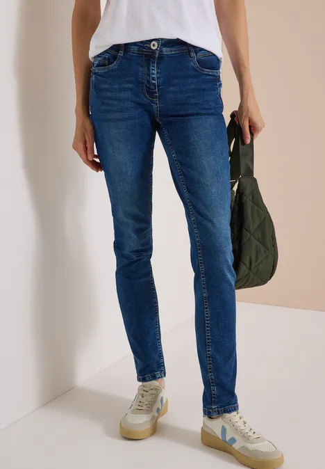 Mid Waist Slim Leg Jeans w stylu Casual Fit mid blue used wash Osoba w obcisłych niebieskich dżinsach, białych trampkach i z pikowaną zieloną torbą.