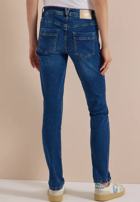 Mid Waist Slim Leg Jeans w stylu Casual Fit mid blue used wash Osoba tyłem w obcisłych niebieskich dżinsach i jasnych sneakersach na neutralnym tle.
