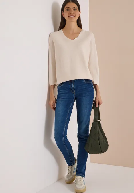 Mid Waist Slim Leg Jeans w stylu Casual Fit mid blue used wash Kobieta w kremowym swetrze z dekoltem w serek, niebieskich jeansach i złotych trampkach, z zieloną torbą.
